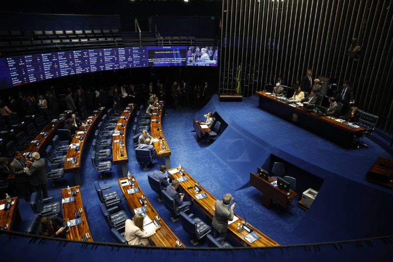 Plenário do Senado Federal com senadores sentados em bancadas curvas de madeira e cadeiras azuis. Painel eletrônico exibe informações legislativas na parede ao fundo. Mesa diretora com cinco pessoas sentadas à direita. Ambiente interno com iluminação artificial e piso azul. Plenário do Senado, em Brasília; senadores negociaram mudanças e aprovaram MP do seguro-defeso - Pedro Ladeira/Folhapress